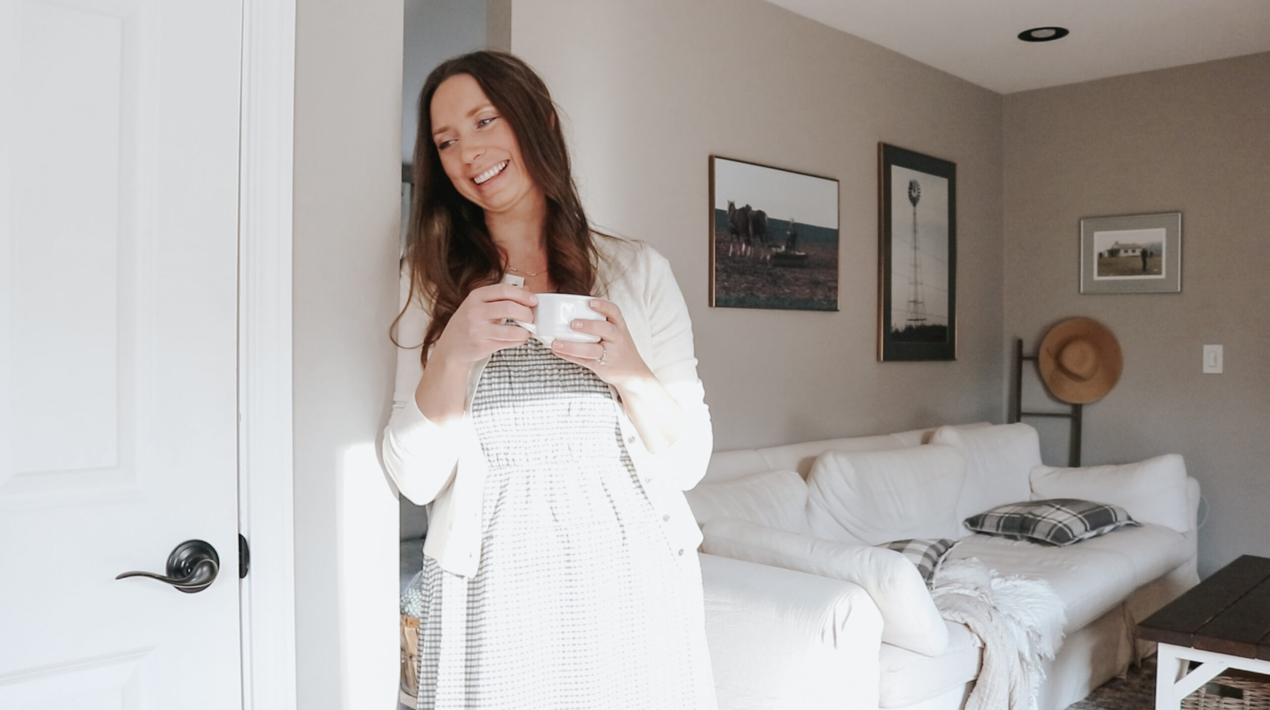 Homemaker leans against a door frame smiling at someone off frame while holding a small white mug. Woman is wearing a dress with a white cardigan with a white couch in the background behind her.